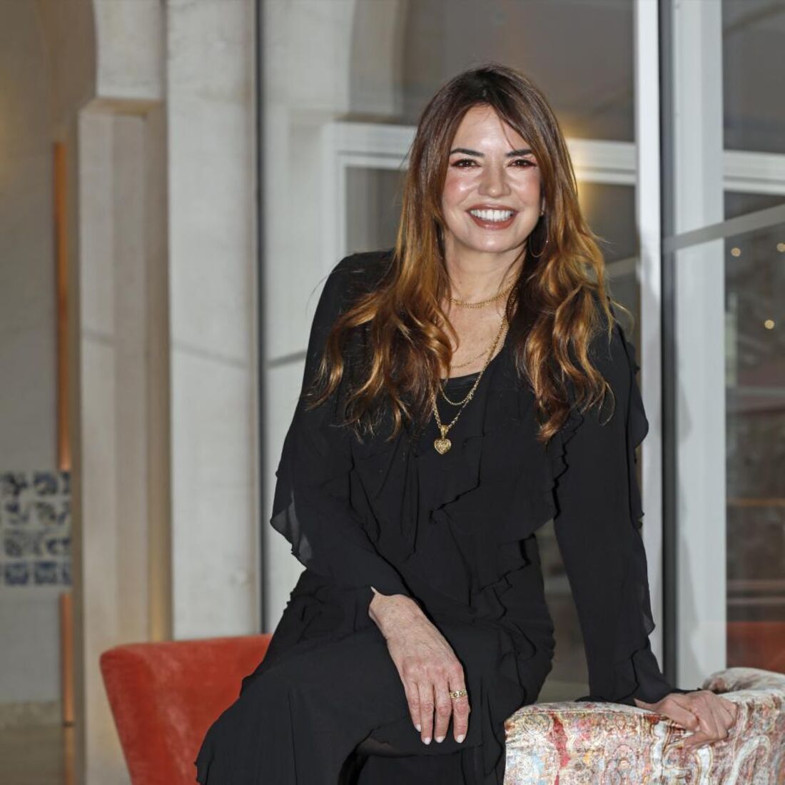 Mulher sorri sentada numa cadeira com um vestido preto e botas