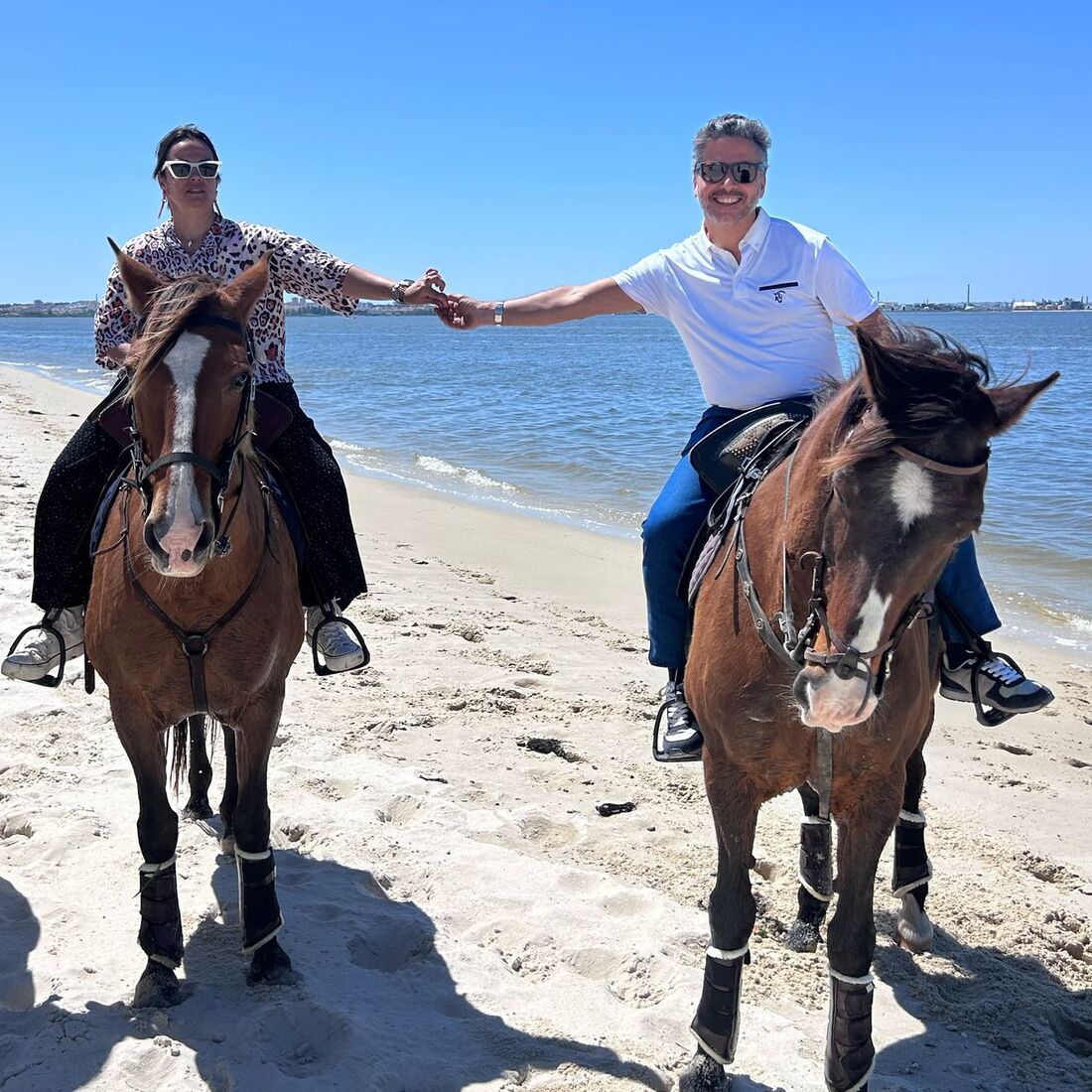 Hugo e Raquel desfrutam de passeio a cavalo na praia sob céu azul.