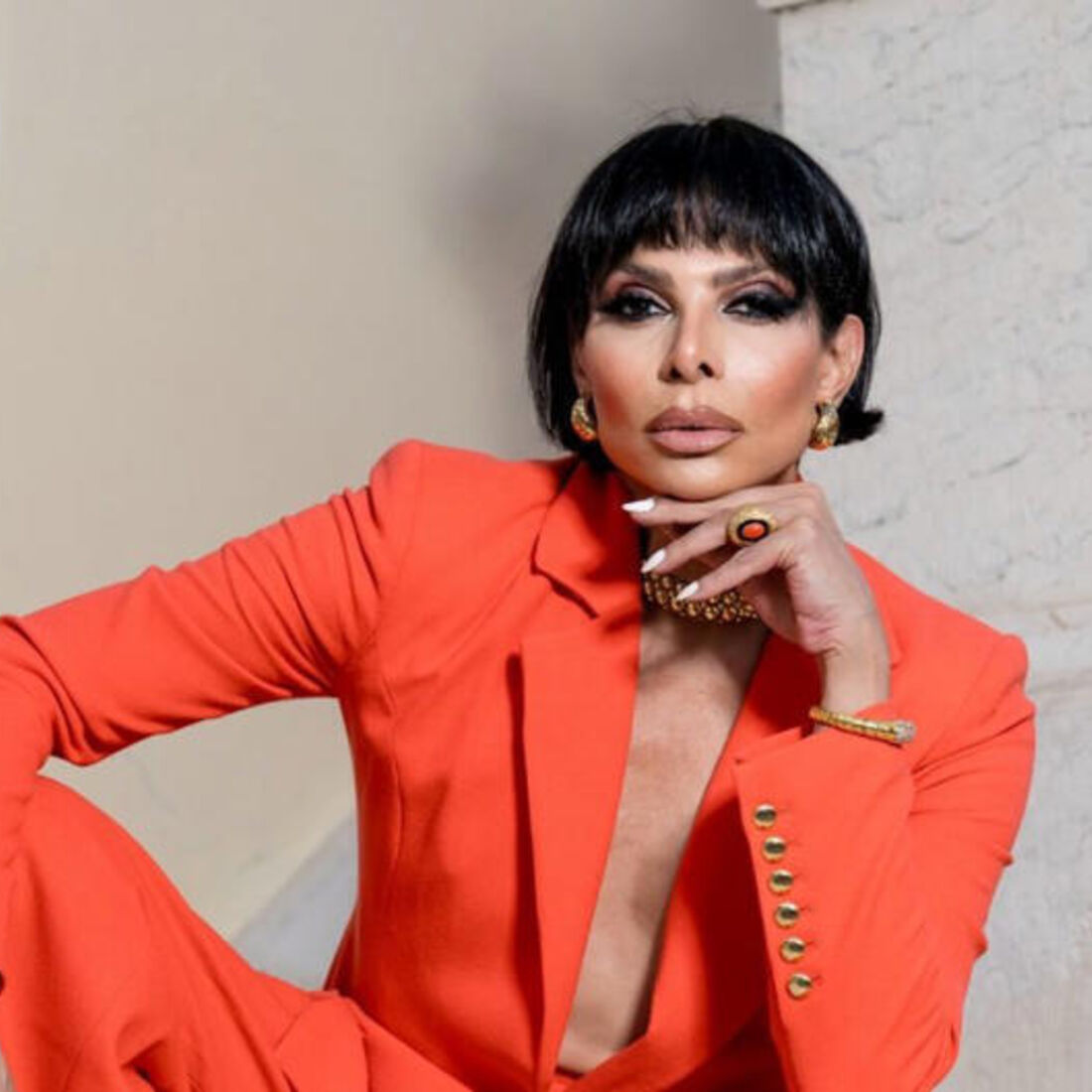 Mulher com fato laranja e cabelo preto exibe joias douradas.
