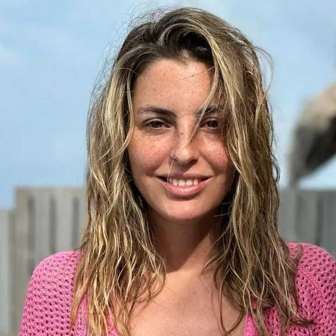 Mulher sorri com um casaco rosa junto ao mar, num dia de sol