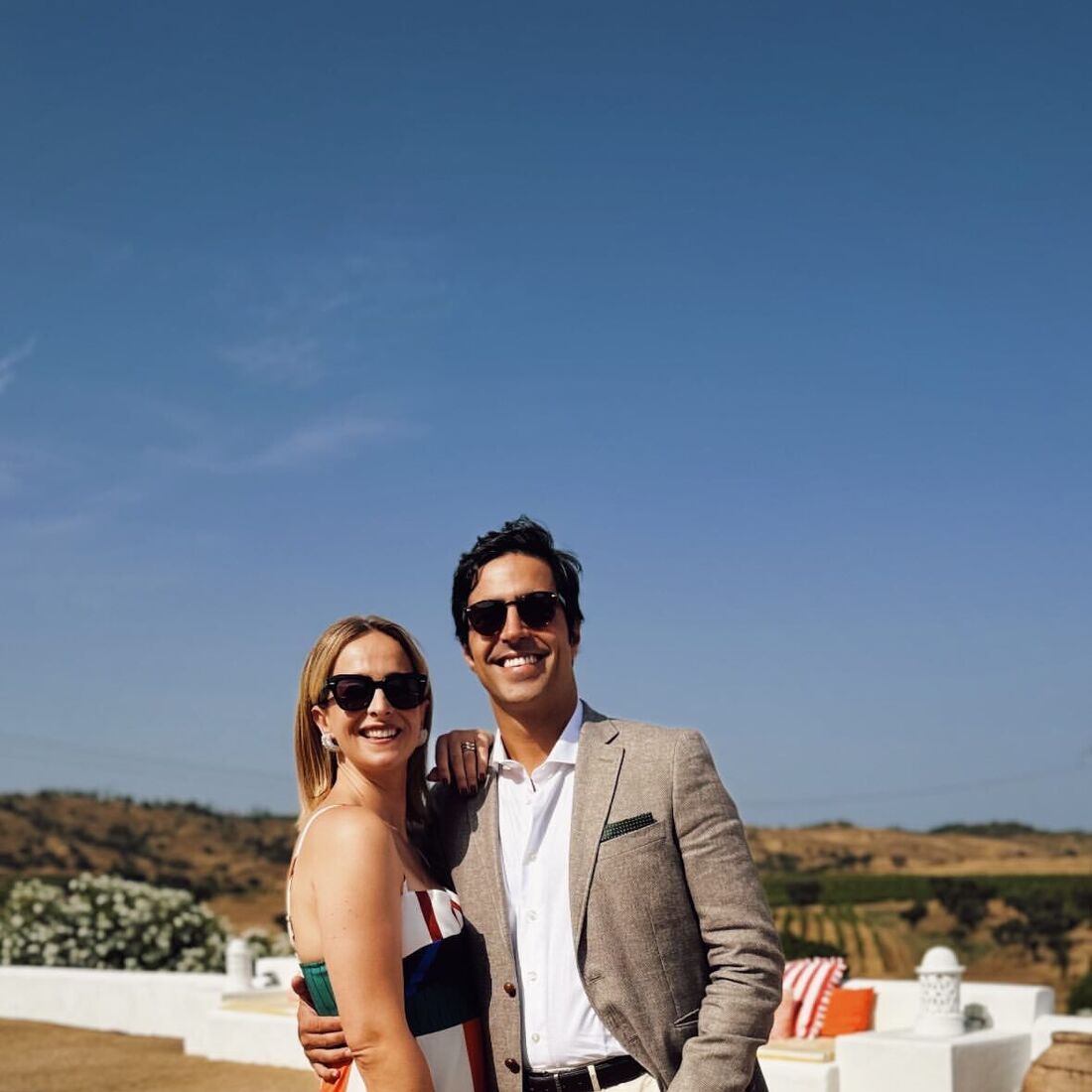 Casal sorridente posa num ambiente campestre; ela veste um vestido às riscas e ele um blazer