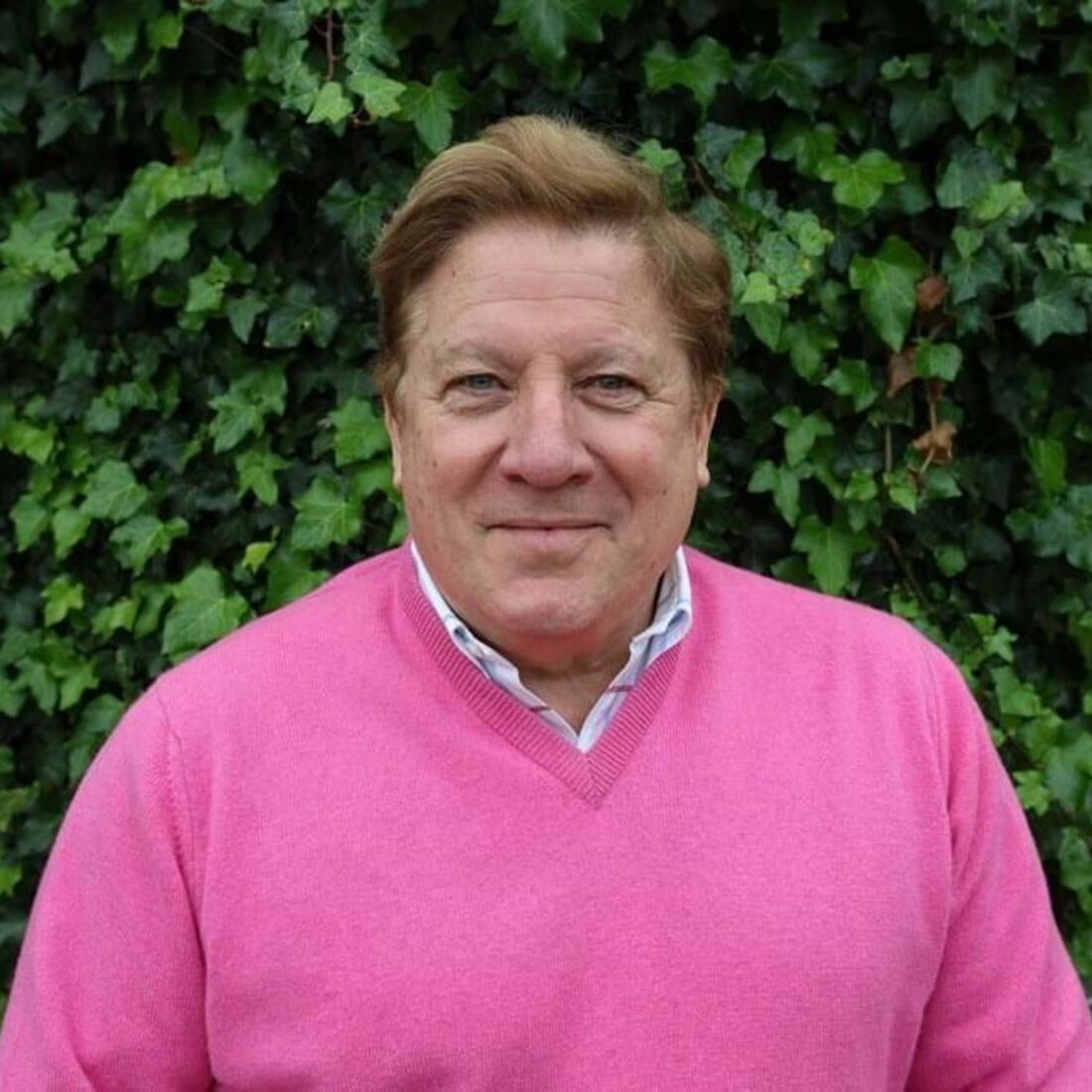 Homem sorri para a câmara, com camisola rosa e fundo de folhagem verde