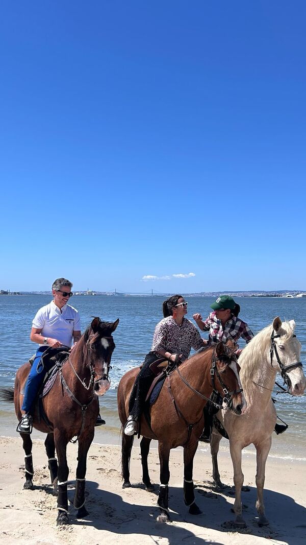 Pessoas andam a cavalo na praia, com o mar e a cidade ao fundo