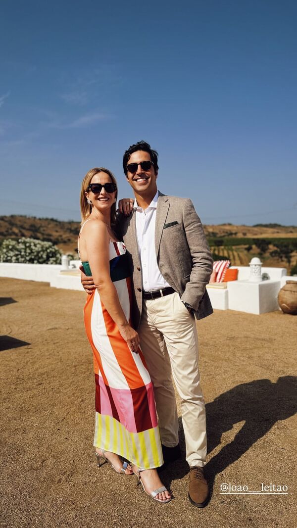 Casal sorridente posa num ambiente campestre; ela veste um vestido às riscas e ele um blazer