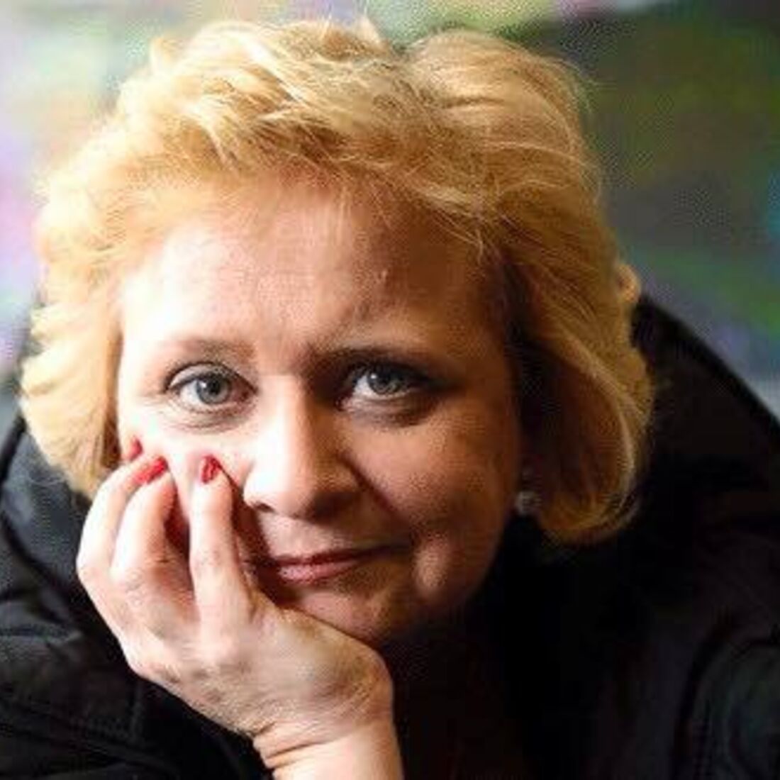 Mulher com cabelo loiro curto e um casaco preto pousa a mão no rosto