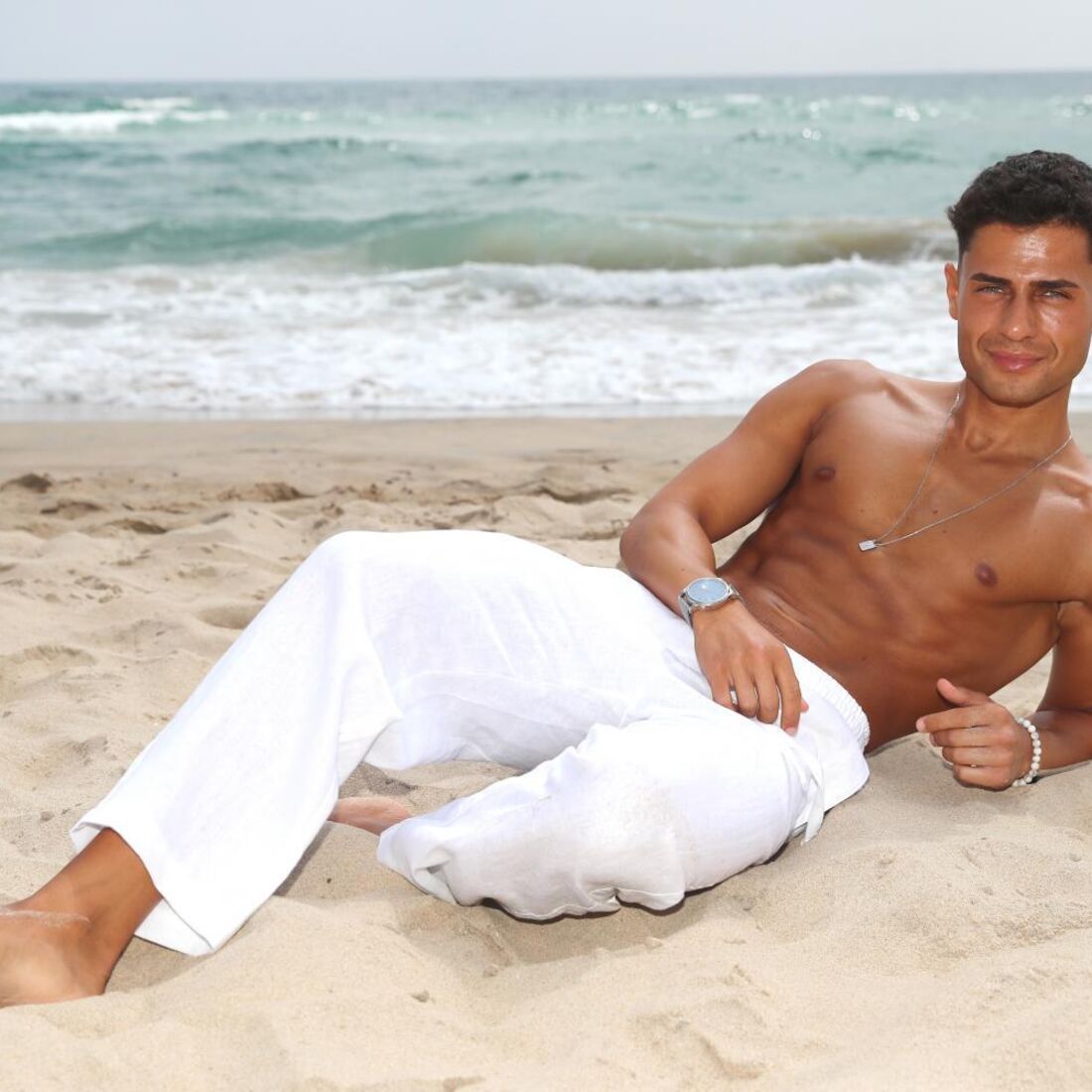 Homem de tronco nu com calças brancas relaxa na praia