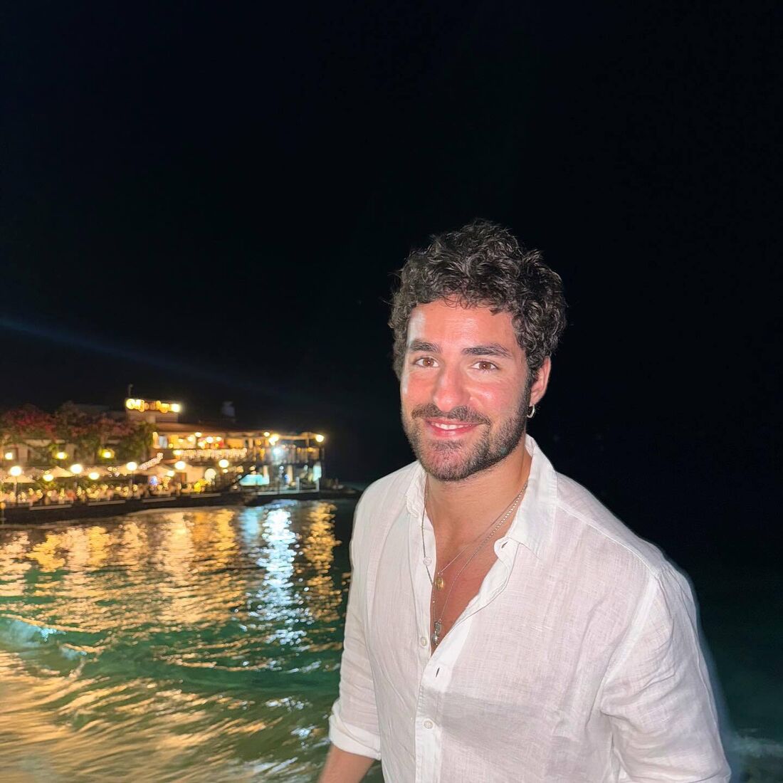 Homem sorri perto da água à noite, com luzes de um edifício ao fundo