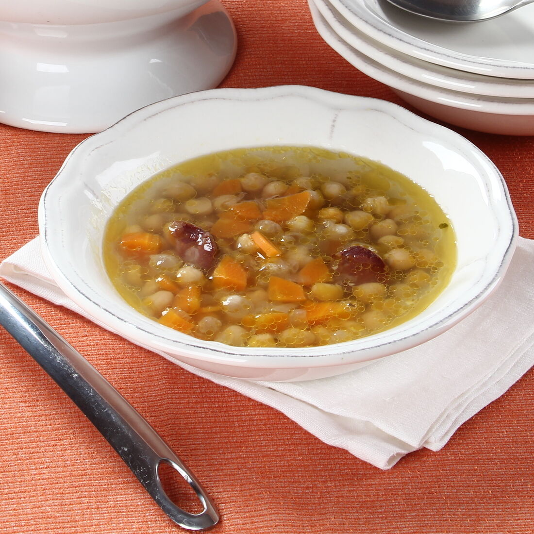 Sopa de grão-de-bico servida com concha e louça branca sobre uma mesa laranja.
